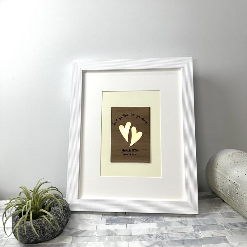 Op de afbeelding: Een rechthoekige houten plaquette met een goudkleurige hartvormige uitsparing, voorzien van de tekst "Loved you then love you always", "Jen & Kim" en "April 13, 2018", geplaatst in een witte lijst met passe-partout en een textuurrand.