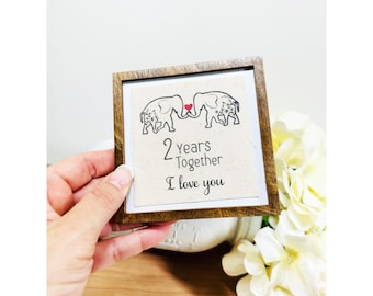 Puede incluir: Un letrero enmarcado en madera con fondo blanco y una ilustración en blanco y negro de dos elefantes con un corazón rojo entre ellos. El texto en el letrero dice "2 años juntos Te amo".
