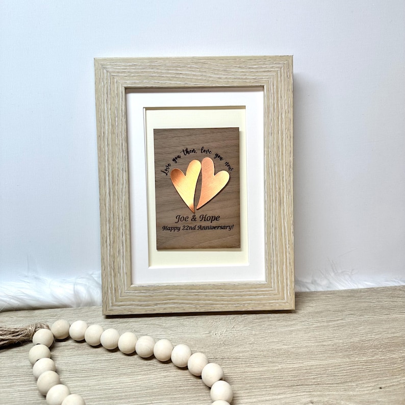 Op de afbeelding: Een lichtgekleurde houten fotolijst met een rechthoekig houten plaatje met twee metalen koperkleurige hartvormige uitsparingen. Het plaatje bevat de tekst: Love you then, love you now, Joe & Hope, Happy 22nd Anniversary, gedrukt in het zwart.