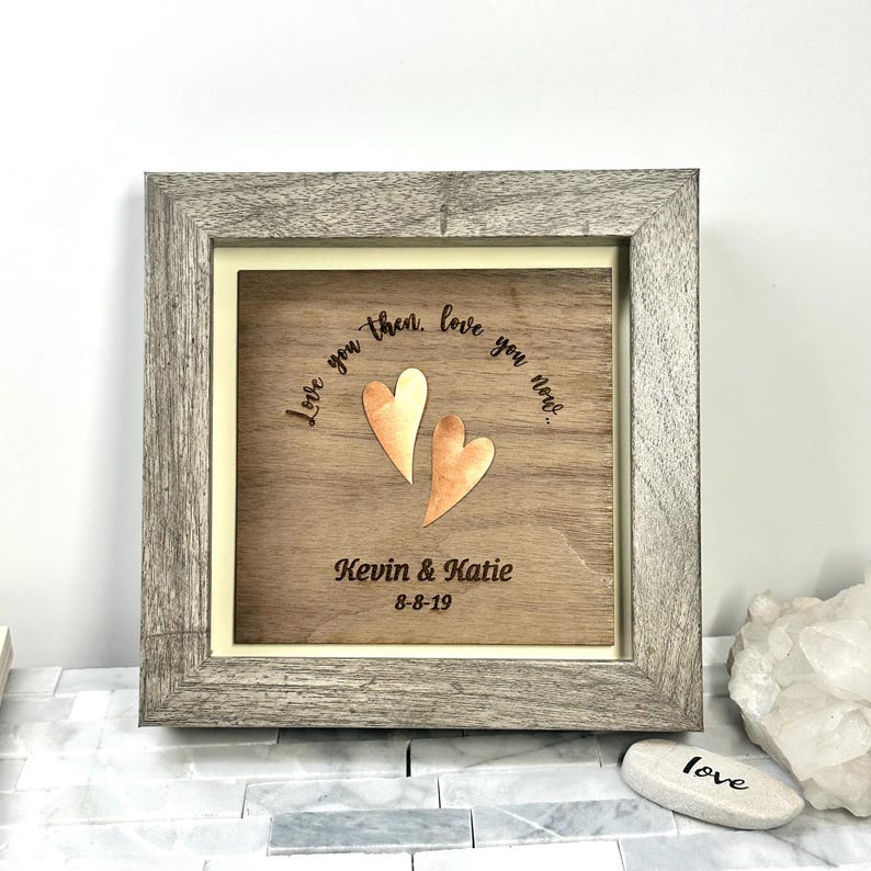 Op de afbeelding: Een vierkante houten lijst met een donkerhouten plaat met twee koperkleurige hartvormige uitsparingen. De tekst "Love you then, love you now..." is boven de harten gegraveerd, met "Kevin & Katie" en "8-8-19" eronder gegraveerd.