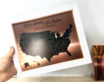 May include: A framed copper map of the United States with a white border. The map is etched with state outlines and includes the text "Life is a Journey Best Traveled Together" and a couple's names. The frame is held by a person.