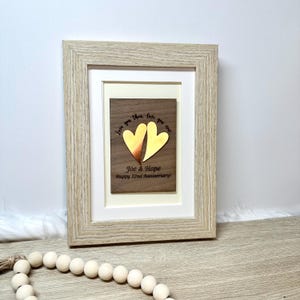 Op de afbeelding: Een licht houten fotolijst met een donkerhouten plaat met twee metalen harten, &eacute;&eacute;n koperkleurig en &eacute;&eacute;n goudkleurig. De tekst luidt: Love you then, love you now, Joe & Hope, Happy 22nd Anniversary! De lijst is voorzien van een witte passe-partout.