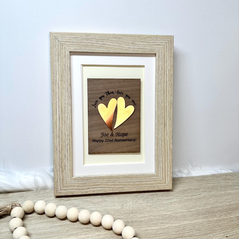 Op de afbeelding: Een licht houten fotolijst met een donkerhouten plaat met twee metalen harten, &eacute;&eacute;n koperkleurig en &eacute;&eacute;n goudkleurig. De tekst luidt: Love you then, love you now, Joe & Hope, Happy 22nd Anniversary! De lijst is voorzien van een witte passe-partout.
