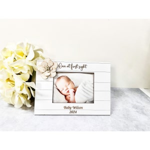 May include: White wooden picture frame with a white flower embellishment. The frame has a photo of a baby inside and the text "Love at first sight" and "Baby Wilson 2024".