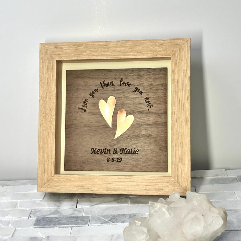 Op de afbeelding: Een vierkante houten schaduwdooslijst met een donkerhouten paneel met twee metallic gouden hartvormige uitsparingen. De tekst "Love you then, love you now..." is erboven gegraveerd, met "Kevin & Katie" en "8-8-19" eronder gedrukt.