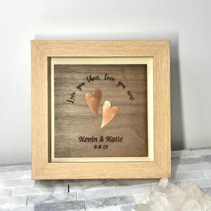 Op de afbeelding: Een vierkante houten lijst met een donkerhouten paneel met twee koperkleurige hartvormige uitsparingen. De tekst "Love you then, love you now..." is boven de harten gegraveerd, met "Kevin & Katie" en "8-8-19" eronder gegraveerd.