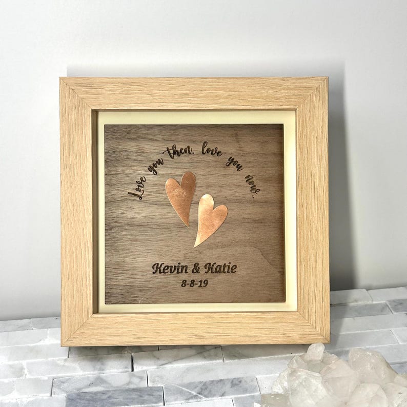 Op de afbeelding: Een vierkante houten lijst met een donkerhouten paneel met twee koperkleurige hartvormige uitsparingen. De tekst "Love you then, love you now..." is boven de harten gegraveerd, met "Kevin & Katie" en "8-8-19" eronder gegraveerd.