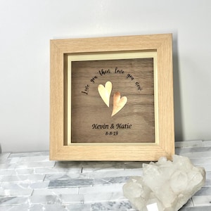 Op de afbeelding: Een vierkante houten lijst met een donkerhouten paneel met twee metalen harten, &eacute;&eacute;n goudkleurig en &eacute;&eacute;n koperkleurig. De tekst "Love you then, love you now..." staat boven de harten, met "Kevin & Katie" en "8-8-19" eronder.