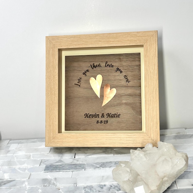 Op de afbeelding: Een vierkante houten lijst met een donkerhouten paneel met twee metalen harten, &eacute;&eacute;n goudkleurig en &eacute;&eacute;n koperkleurig. De tekst "Love you then, love you now..." staat boven de harten, met "Kevin & Katie" en "8-8-19" eronder.