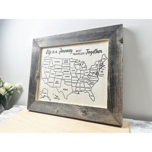 May include: A framed map of the United States printed on a light-colored fabric background. The map features black outlines of state borders and names. Above the map, the text reads, Life is a Journey BEST TRAVELED Together. The frame is dark wood.
