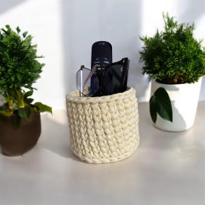 May include: A white crocheted basket with a remote control, sunglasses, and eyeglasses inside. The basket is sitting on a white surface.