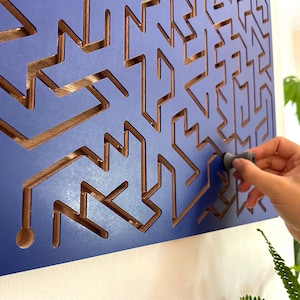 May include: A blue wooden wall art piece with a geometric design. The design features cut-out shapes revealing a wood grain interior. A hand is shown interacting with a small, grey, cylindrical object within the artwork.
