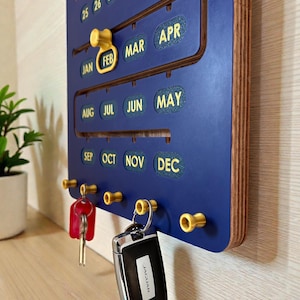 May include: A blue wooden calendar with gold accents. The calendar displays months and dates, with a gold pin marking February. Key hooks hold keys and a car key fob. The car key fob has the word "JAGUAR" on it.