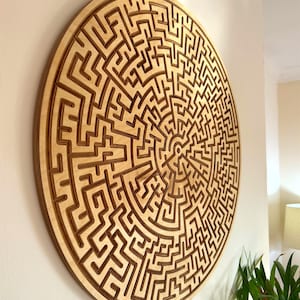May include: A large, round wooden wall art piece featuring a complex maze design. The maze is carved into the light-colored wood, creating a contrast with the darker lines of the maze. The artwork is mounted on a white wall.