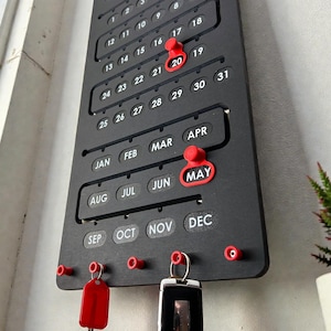 May include: A black wall calendar with red accents, featuring dates and months. Red push pins mark specific dates, with a key holder at the bottom. The month of May is highlighted. Keys hang from the key holder.