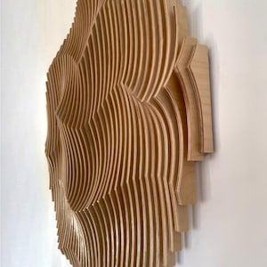 May include: A three-dimensional wooden wall art piece. The artwork is constructed from numerous thin, curved wooden strips, creating a layered, wave-like design. The natural wood color and the interplay of light and shadow give it a modern aesthetic.