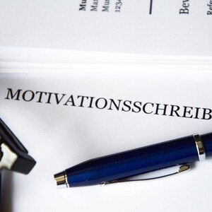 以下が含まれることがあります： ドイツ語の「MOTIVATIONSSCHREIBEN」という文字が黒で印刷された書類のクローズアップ。青いペンと眼鏡が書類の上にあります。背景には白い紙の束があります。