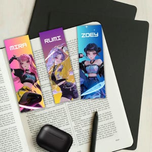 May include: Three colorful bookmarks featuring anime-style illustrations of female characters. Each bookmark has a different name: "MIRA", "RUMI", and "ZOEY". The bookmarks are on a book with a black pen and a black wireless earbud case.