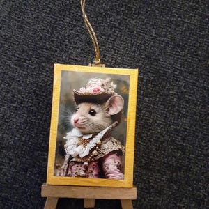 May include: A decorative ornament featuring a framed image of a mouse dressed in ornate clothing and a hat. The image is set on a miniature wooden easel and has a gold-colored frame. A gold string is attached for hanging.