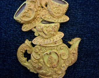 May include: A yellow decorative ornament featuring a stack of three teacups resting atop a teapot. The object has a textured, ornate surface with raised relief details and is suspended by a thin metallic cord attached to the top teacup.