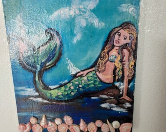 May include: Hand-painted mermaid art on a wooden plaque. The mermaid has long blonde hair, a pink torso, and a green tail. The background is blue with white floral accents. Seashells are glued to the bottom edge. A blue string is attached for hanging.