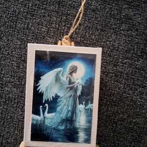 May include: A small, rectangular canvas print featuring a serene scene of an angel with white wings standing near swans in a moonlit lake. The print is mounted on a miniature wooden easel and has a gold string for hanging.