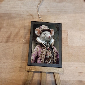 May include: A small wooden easel displays a framed image of a mouse dressed in ornate, regal attire. The mouse wears a hat and a pink and gold jacket. A gold string is attached to the top of the frame for hanging. The easel is made of light brown wood.