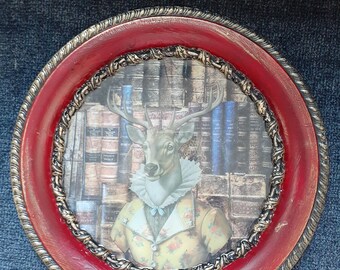 May include: A round framed picture featuring a deer with antlers dressed in a yellow floral jacket and a ruffled collar. The background shows a library of books. The frame is red with a gold rope-like trim.