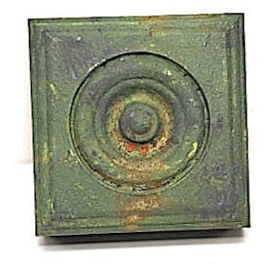 May include: A square, green metal plaque with a circular design in the center. The circle has concentric rings of red, yellow, and black paint, with a small, round, black knob in the center.