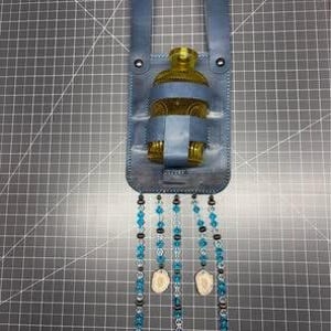 May include: A blue leather holder containing a small amber glass bottle. The holder features two vertical straps at the top and five dangling strands decorated with blue and clear beads, dark spacers, and two light-colored, flat, oval-shaped charms.