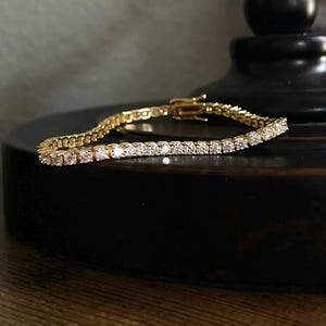 May include: A gold-toned tennis bracelet featuring a continuous row of clear, round-cut stones set in a metallic frame. The bracelet includes a secure box clasp mechanism and is displayed in a curved shape, showcasing the reflective quality of the stones.
