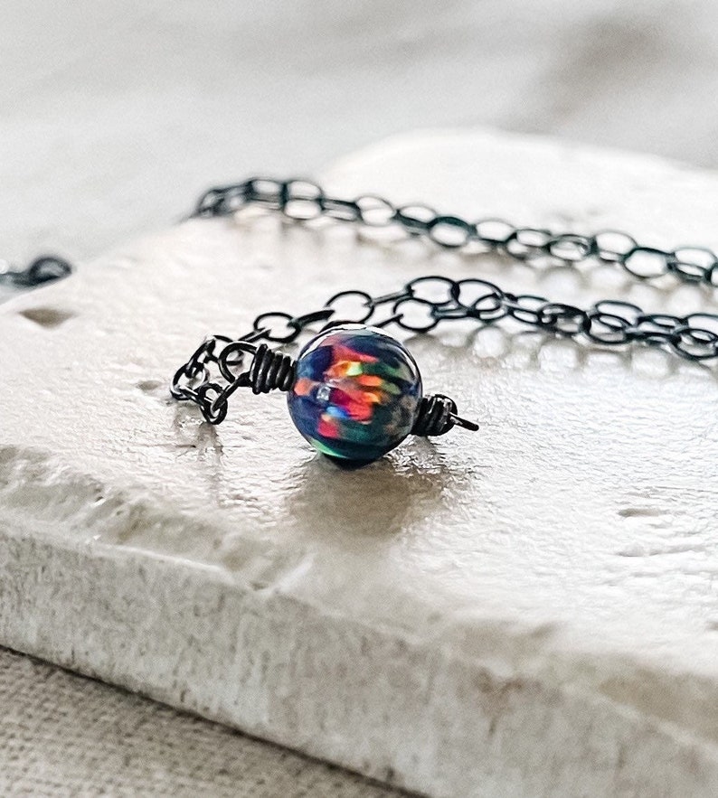 Pu&ograve; includere: Una collana a catena nera con un unico ciondolo di opale iridescente. Il ciondolo &egrave; avvolto in filo nero e presenta un arcobaleno di colori.