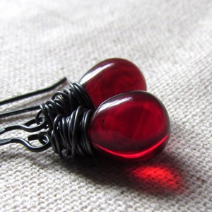 Pode incluir: Um par de brincos com contas de vidro vermelho em forma de lágrima e fio preto enrolado em torno das contas.