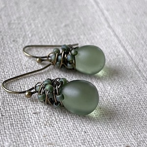 Pode incluir: Um par de brincos pendentes com contas em forma de gota de cor verde-s&aacute;lvia fosca e lisa. Cada conta &eacute; envolvida em fio de bronze escuro e decorada com pequenas contas de semente a condizer. Os brincos pendem de ganchos simples de metal cor de bronze.