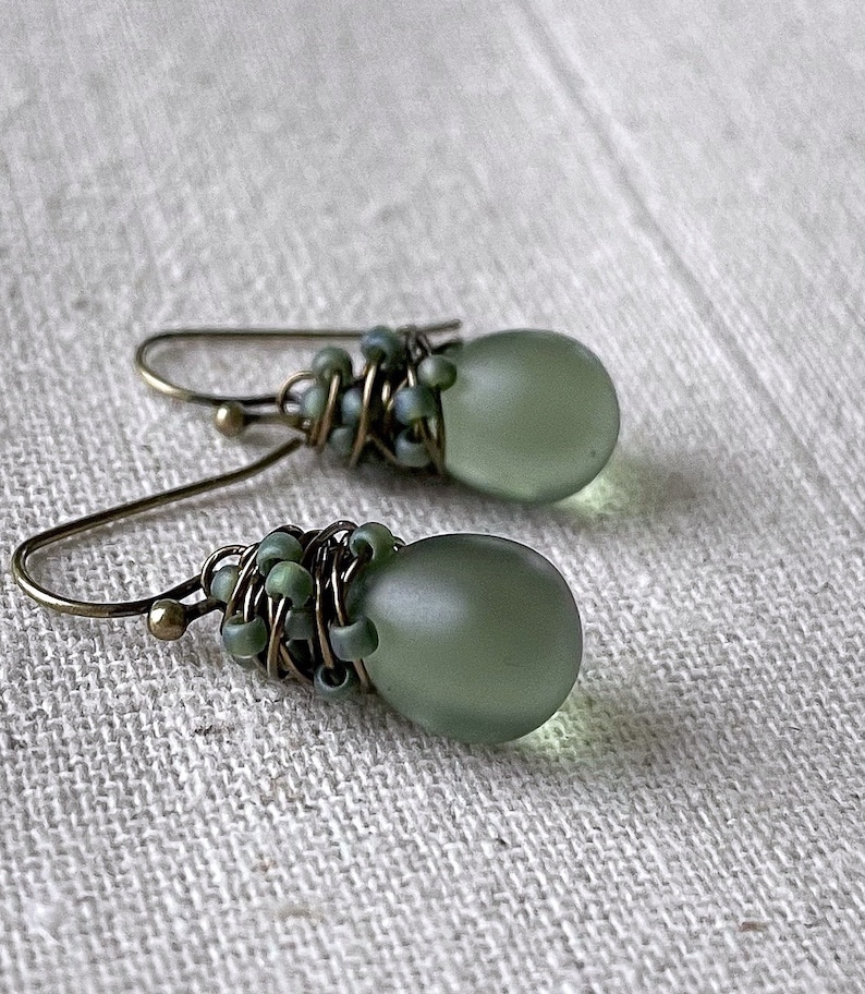 Pode incluir: Um par de brincos pendentes com contas em forma de gota de cor verde-s&aacute;lvia fosca e lisa. Cada conta &eacute; envolvida em fio de bronze escuro e decorada com pequenas contas de semente a condizer. Os brincos pendem de ganchos simples de metal cor de bronze.