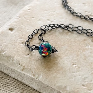 Pu&ograve; includere: Una delicata collana a catena nera con un unico ciondolo di opale iridescente. Il ciondolo &egrave; avvolto da un filo nero e presenta un arcobaleno di colori.