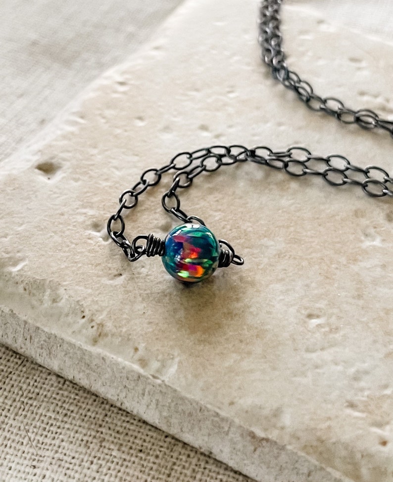Pu&ograve; includere: Una delicata collana a catena nera con un unico ciondolo di opale iridescente. Il ciondolo &egrave; avvolto da un filo nero e presenta un arcobaleno di colori.