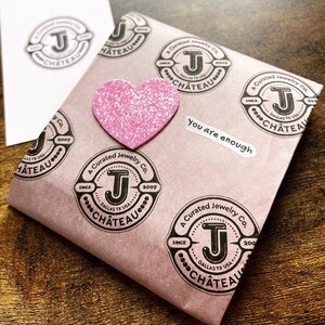 May include: A pink paper bag with a pink glitter heart sticker and multiple black and white stamps that say "A Curated Jewelry Co. Since 2009 Dallas TX USA Chateau". The bag also has a white sticker that says "You are enough".