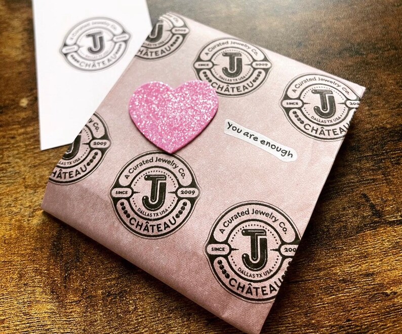 May include: A pink paper bag with a pink glitter heart sticker and multiple black and white stamps that say "A Curated Jewelry Co. Since 2009 Dallas TX USA Chateau". The bag also has a white sticker that says "You are enough".