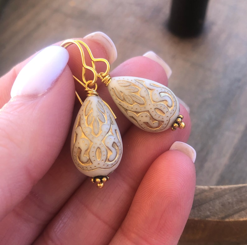 May include: A pair of gold dangle earrings with white teardrop-shaped beads. The beads have a gold filigree design.
