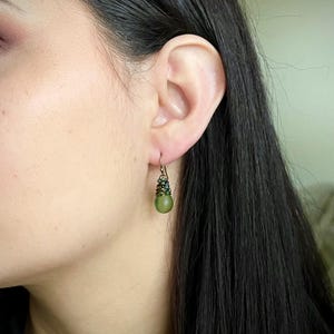 Pode incluir: Um brinco pendente com uma conta esf&eacute;rica verde-oliva mate suspensa por um gancho de arame cor de bronze. O topo da conta &eacute; envolvido por arame escuro e pequenas contas facetadas verde-escuras, criando um efeito texturizado e em camadas.