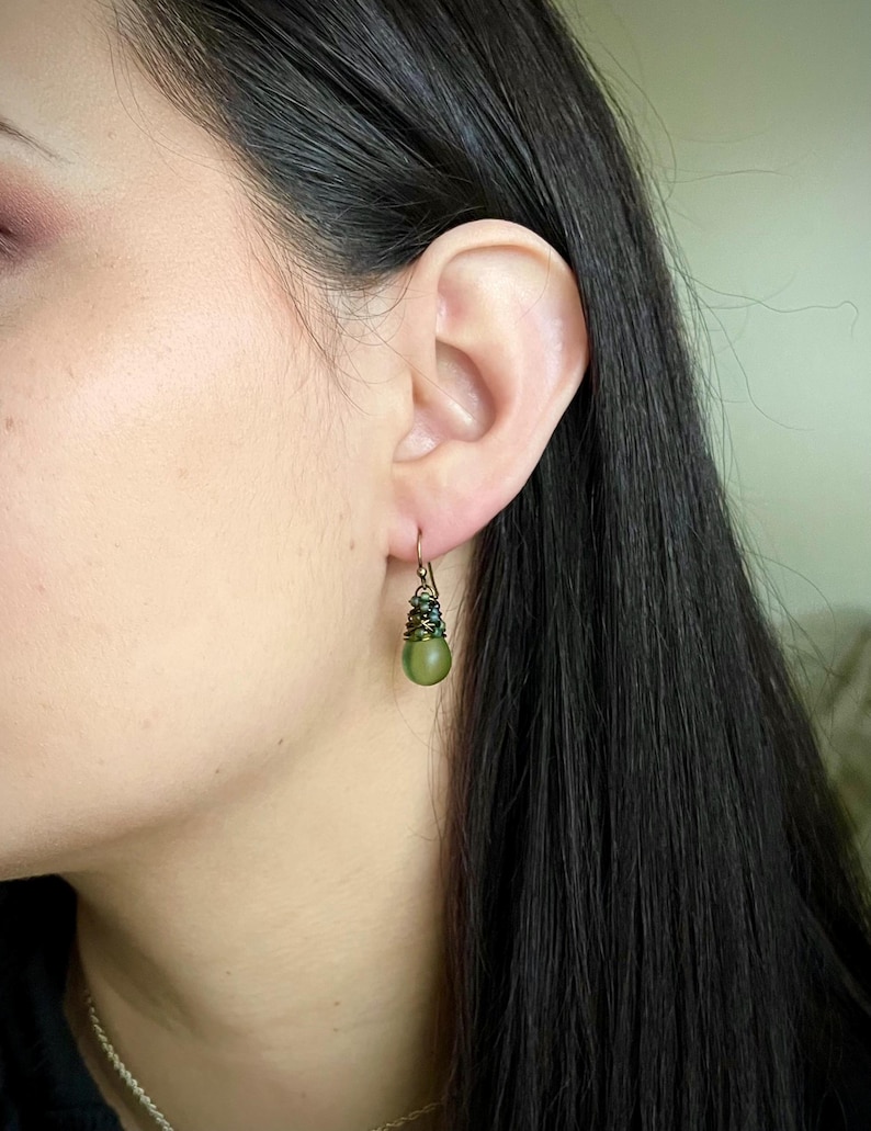 Pode incluir: Um brinco pendente com uma conta esf&eacute;rica verde-oliva mate suspensa por um gancho de arame cor de bronze. O topo da conta &eacute; envolvido por arame escuro e pequenas contas facetadas verde-escuras, criando um efeito texturizado e em camadas.