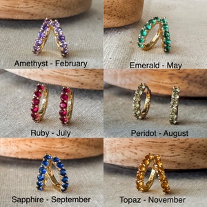 May include: Six pairs of gold hoop earrings with different colored gemstones. Each pair is labeled with the gemstone name and corresponding birth month. The gemstones are amethyst, emerald, ruby, peridot, sapphire, and topaz.