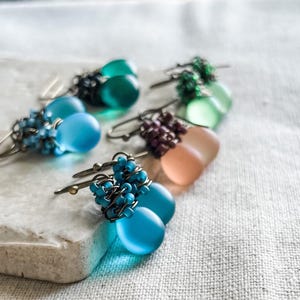 Pode incluir: Uma cole&ccedil;&atilde;o de brincos pendentes com contas de vidro fosco em forma de l&aacute;grima em tons de azul, verde e p&ecirc;ssego. Cada brinco &eacute; acentuado com grupos de pequenas contas a condizer, enroladas em fio de metal escuro com ganchos.
