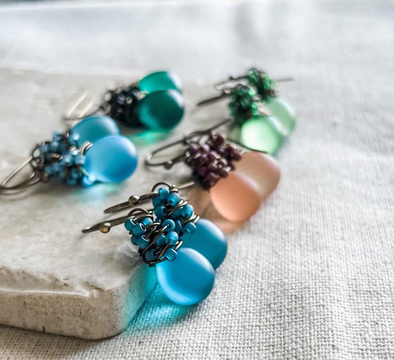 Pode incluir: Uma cole&ccedil;&atilde;o de brincos pendentes com contas de vidro fosco em forma de l&aacute;grima em tons de azul, verde e p&ecirc;ssego. Cada brinco &eacute; acentuado com grupos de pequenas contas a condizer, enroladas em fio de metal escuro com ganchos.