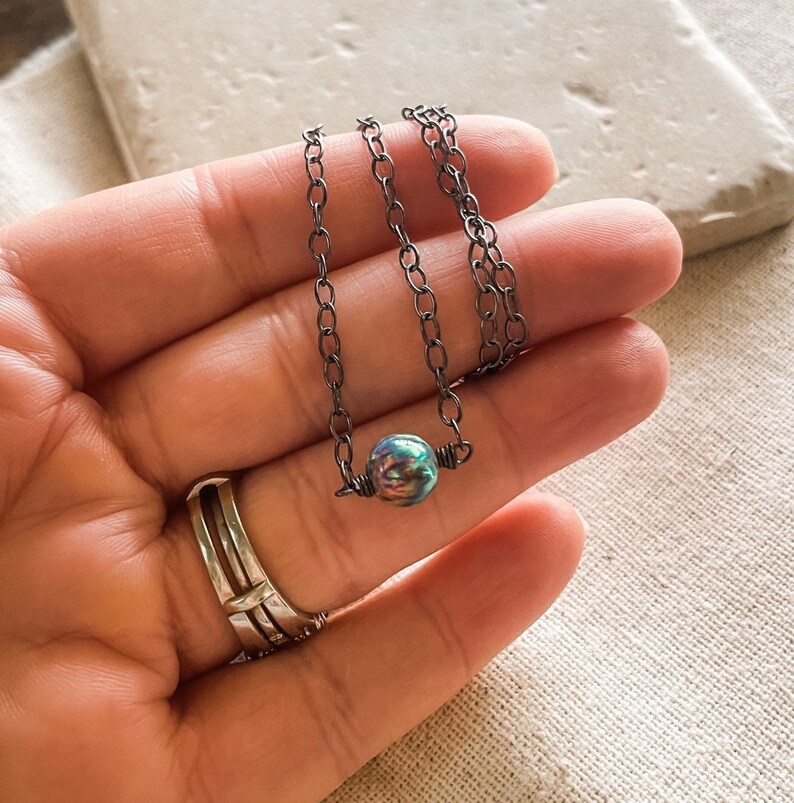 Pu&ograve; includere: Una delicata collana a catena d'argento con un ciondolo a forma di perla iridescente. La perla presenta un motivo unico e vorticoso di blu, verde e viola.