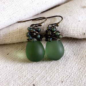 May include: A pair of green glass teardrop earrings with bronze wire wrapped around the top of each earring. The earrings are on a white fabric background.