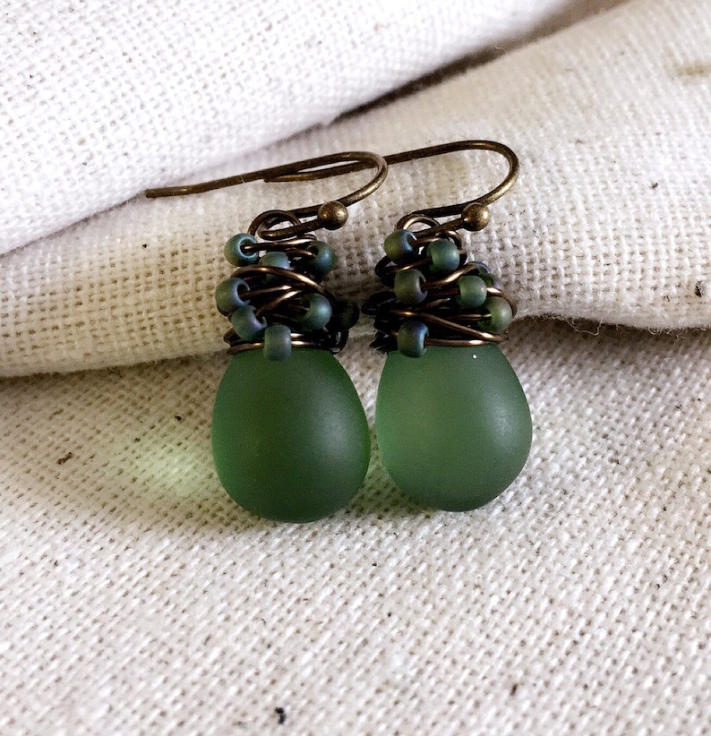 Pode incluir: Um par de brincos pendentes com contas em forma de gota verde-s&aacute;lvia com acabamento mate, suspensas por ganchos de arame cor de bronze. A parte superior &eacute; envolvida por arame de bronze escuro com pequenas contas a condizer.