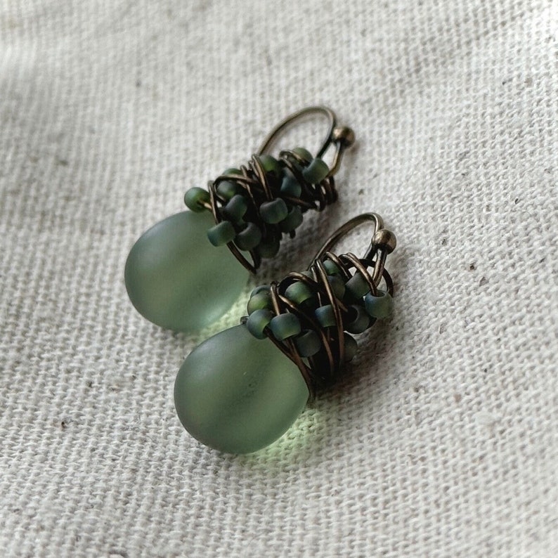 Pode incluir: Um par de brincos pendentes com contas de vidro fosco verde-s&aacute;lvia em forma de l&aacute;grima. O topo de cada conta &eacute; envolvido por fio de tom bronze escuro e acentuado com pequenas contas de semente verde mate, finalizado com um gancho simples.
