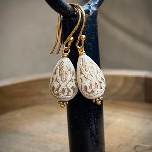 May include: A pair of gold dangle earrings with white teardrop-shaped beads. The beads have a delicate, intricate design etched into them.